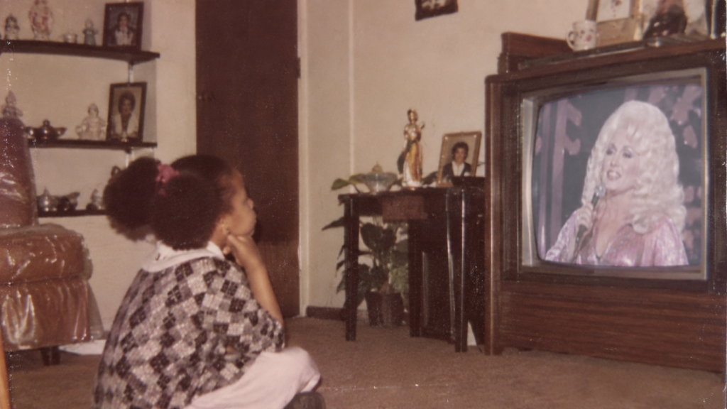 Young Hedreich Nichols with afro puffs watching Dolly Parton perform on television in a 1970s family living room.
